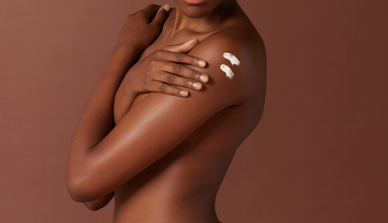 Une femme noire aux épaules nues croise les bras sur sa poitrine. Deux petites touches de crème blanche sont appliquées sur son épaule.