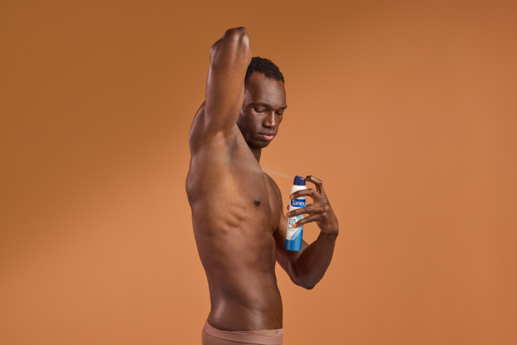 Un homme noir au torse nu lève le bras pour s'appliquer un déodorant en aérosol Sanex sur son aisselle. Il est de profil sur un fond ocre uni.