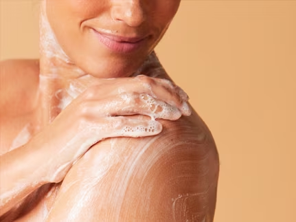 Gros plan sur une femme souriante qui savonne délicatement son épaule et sa clavicule avec une mousse onctueuse.