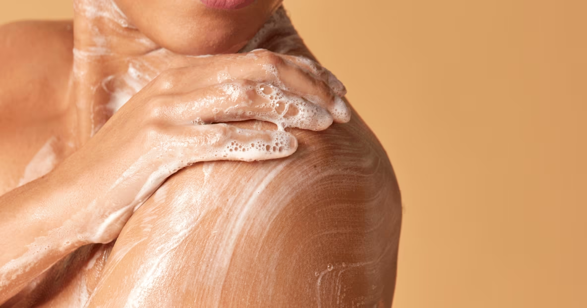Gros plan sur une femme souriante qui savonne délicatement son épaule et sa clavicule avec une mousse onctueuse.