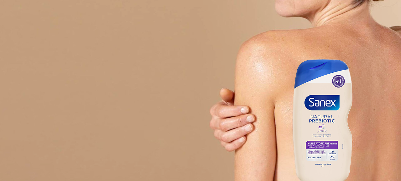Bannière publicitaire montrant une femme de dos, sa main posée sur son épaule. Un flacon d'huile lavante Sanex Natural Prebiotic Atopicare Repair est superposé sur l'image.