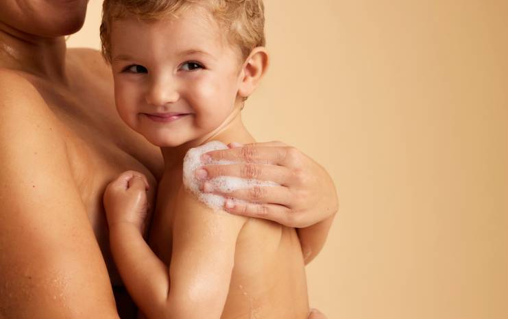Un jeune enfant aux cheveux blonds regarde la caméra avec un léger sourire, tandis qu'un adulte lave tendrement son épaule avec une main pleine de mousse.