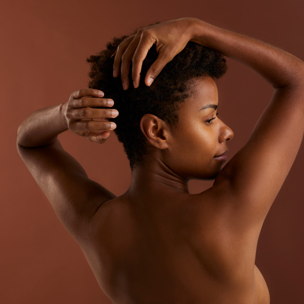 Vue de dos d'une femme noire aux cheveux courts, ses mains posées sur sa nuque, sur un fond marron uni.
