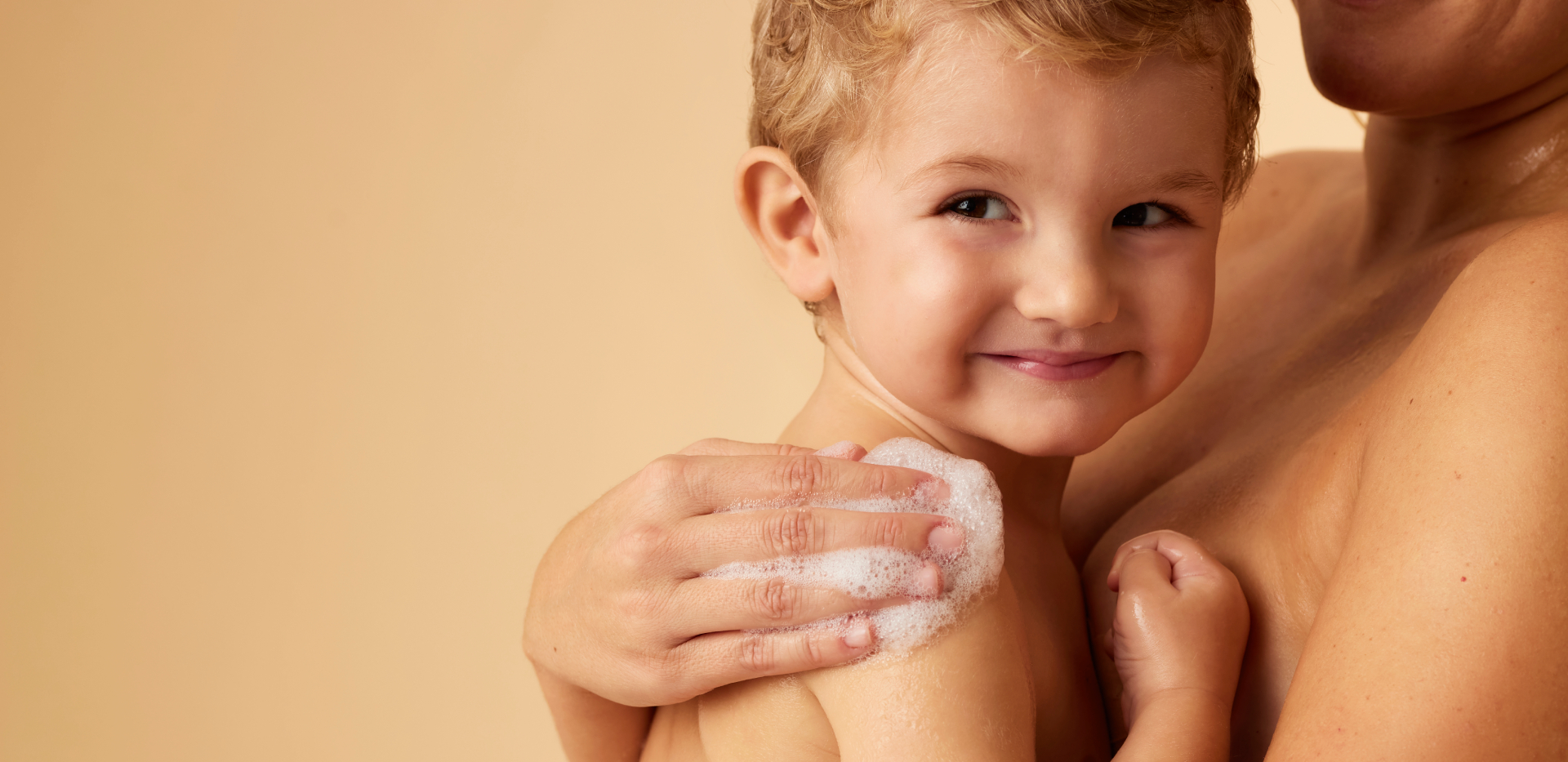 Un jeune enfant aux cheveux blonds regarde la caméra avec un léger sourire, tandis qu'un adulte lave tendrement son épaule avec une main pleine de mousse.